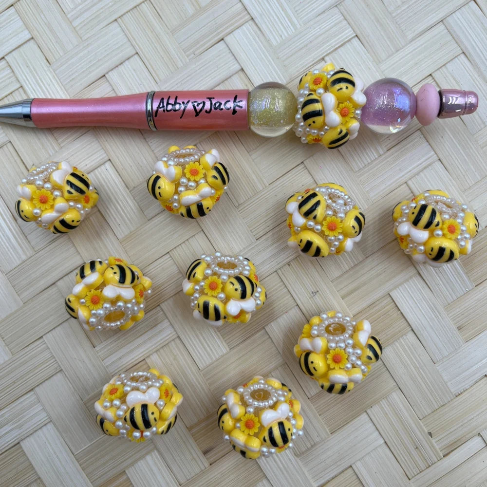 Chunky yellow and black bumble bee fancy beads decorated with white pearl accents, sunflowers, and bee figures, displayed beside a rose gold beadable pen on a woven surface.