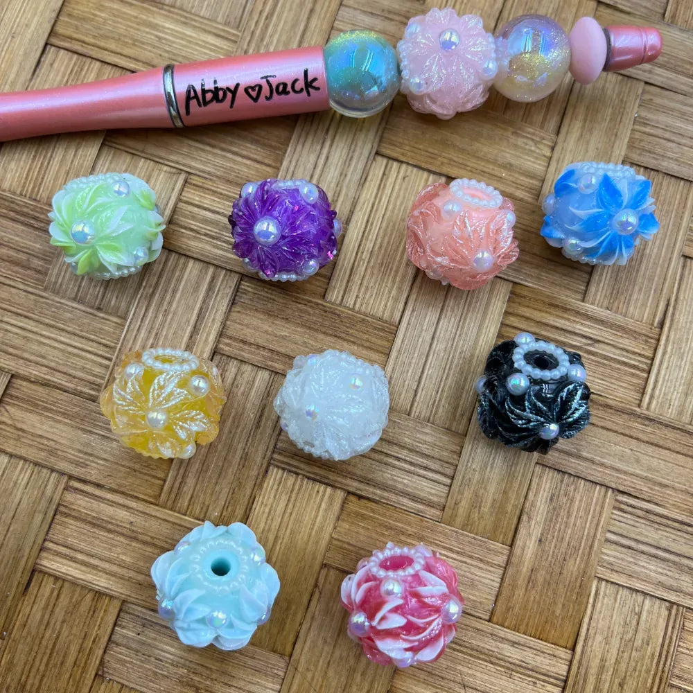 Assorted floral carved chunky resin beads in multiple colors including purple, pink, blue, green, yellow, white, and black with iridescent pearl accents, displayed on a woven surface next to a pink beadable pen labeled Abby Jack.