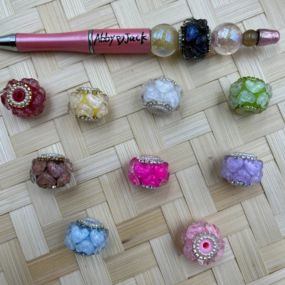 Assorted chunky floral textured fancy beads with rhinestone and gold/silver trim edges in red, yellow, white, green, mauve, hot pink, lavender, blue, and pink, displayed beside a pink beadable pen on a wicker surface.