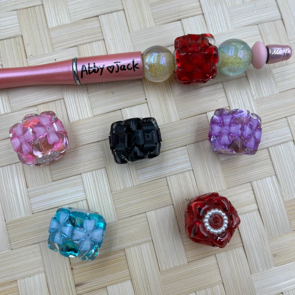Chunky square flower cluster acrylic fancy beads in assorted colors including pink, black, purple, teal, and red displayed on a woven bamboo mat next to a beadable pen labeled Abby and Jack.