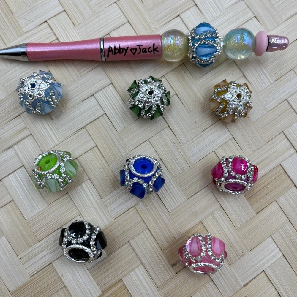 Assorted rhinestone crystal fancy chunky beads in blue, green, pink, black, and gold with silver metal accents, displayed next to a pink beadable pen on a woven surface.