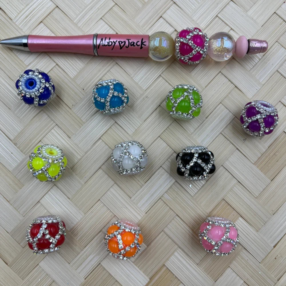 Assorted rhinestone encrusted chunky fancy beads in multiple colors including pink, teal, green, purple, yellow, white, black, red, orange, and blue evil eye, displayed on a woven bamboo surface next to a pink beadable pen branded Abby Jack.