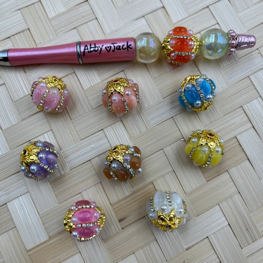 Assorted rhinestone gold filigree chunky fancy beads in pink, lavender, peach, blue, yellow, white, and orange displayed on a wicker surface next to a decorated beadable pen labeled Abby Jack.