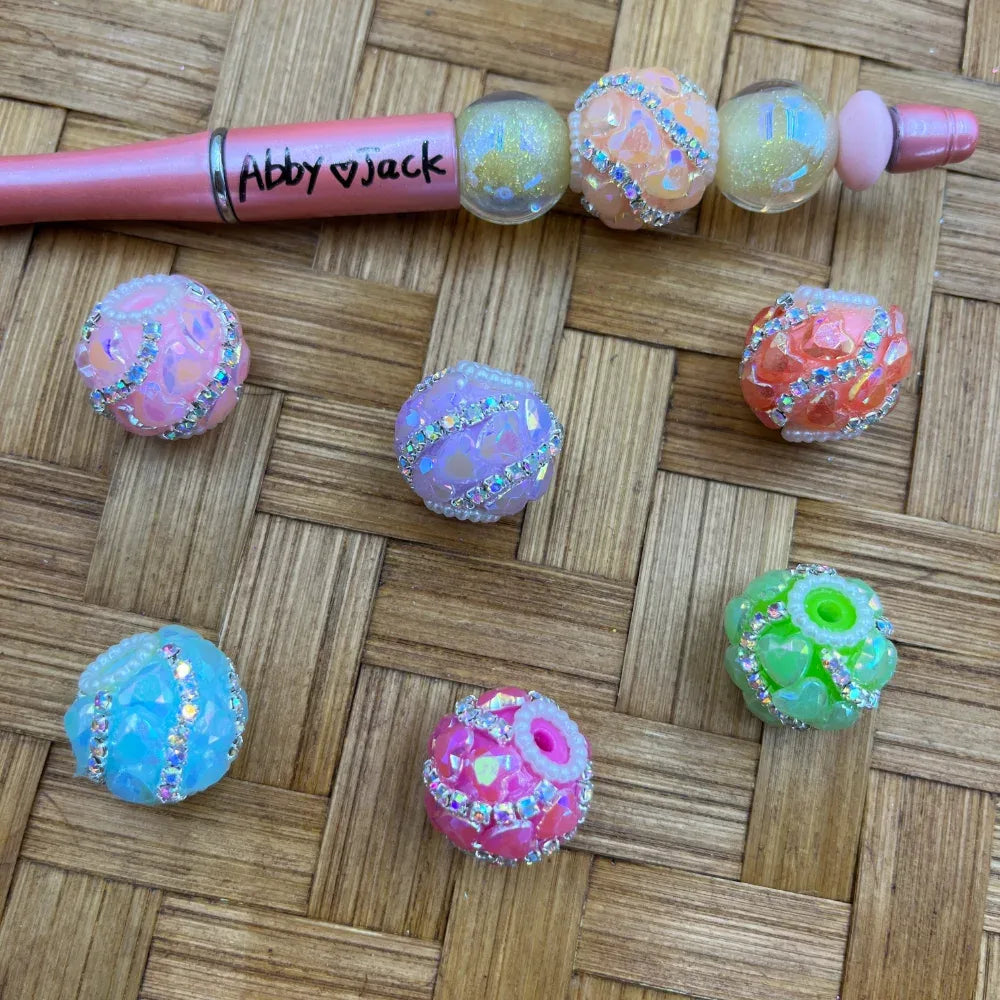 Assorted chunky holographic resin fancy beads with rhinestone bands in pastel pink, purple, blue, orange, green, and white, displayed on a woven surface alongside a pink beadable pen labeled Abby and Jack.