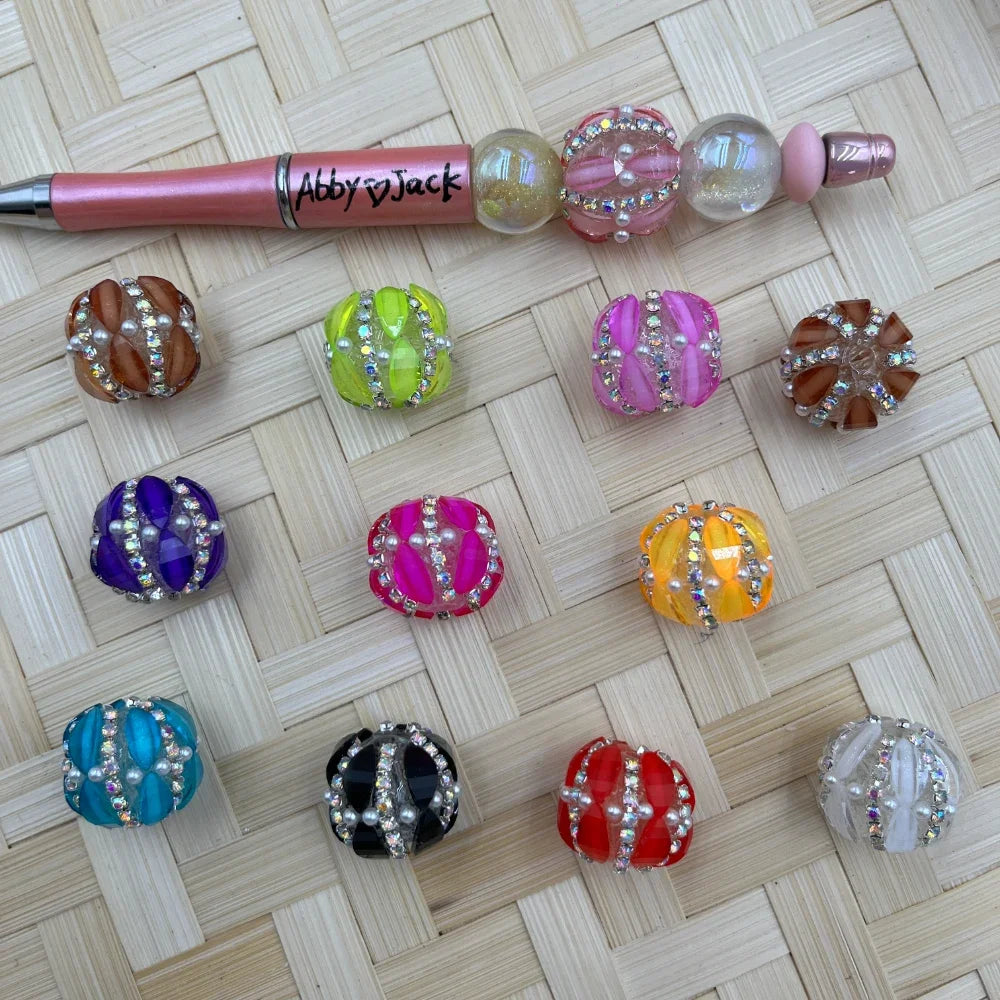 Assorted rhinestone and pearl embellished fancy chunky beads in 11 colors including purple, pink, orange, red, black, teal, green, brown, and white, displayed next to a beadable pen on a woven surface.