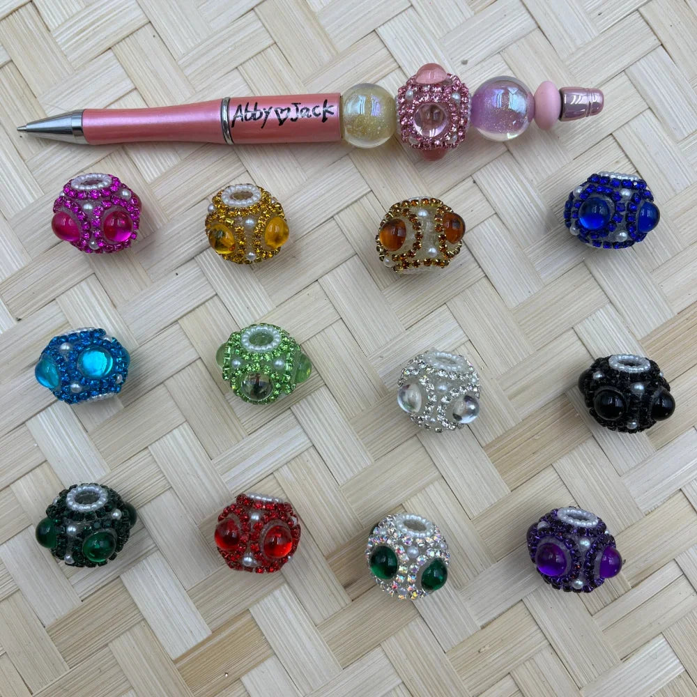 Assorted rhinestone and pearl chunky fancy beads in 12 colors including pink, yellow, blue, green, red, purple, black, and clear, displayed next to a beadable pen on a woven bamboo surface.