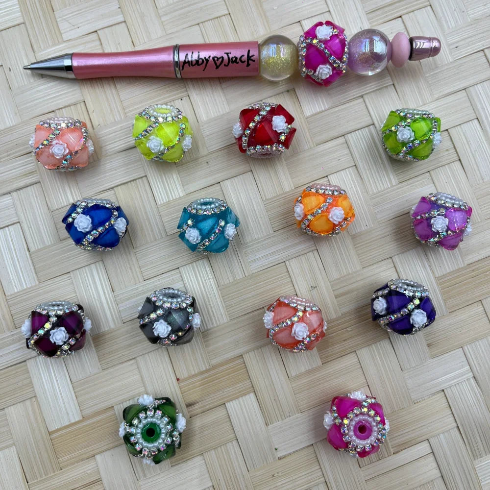 Assorted colorful chunky fancy beads decorated with rhinestones and white rose flowers, displayed on woven surface alongside a pink beadable pen labeled Abby Jack.
