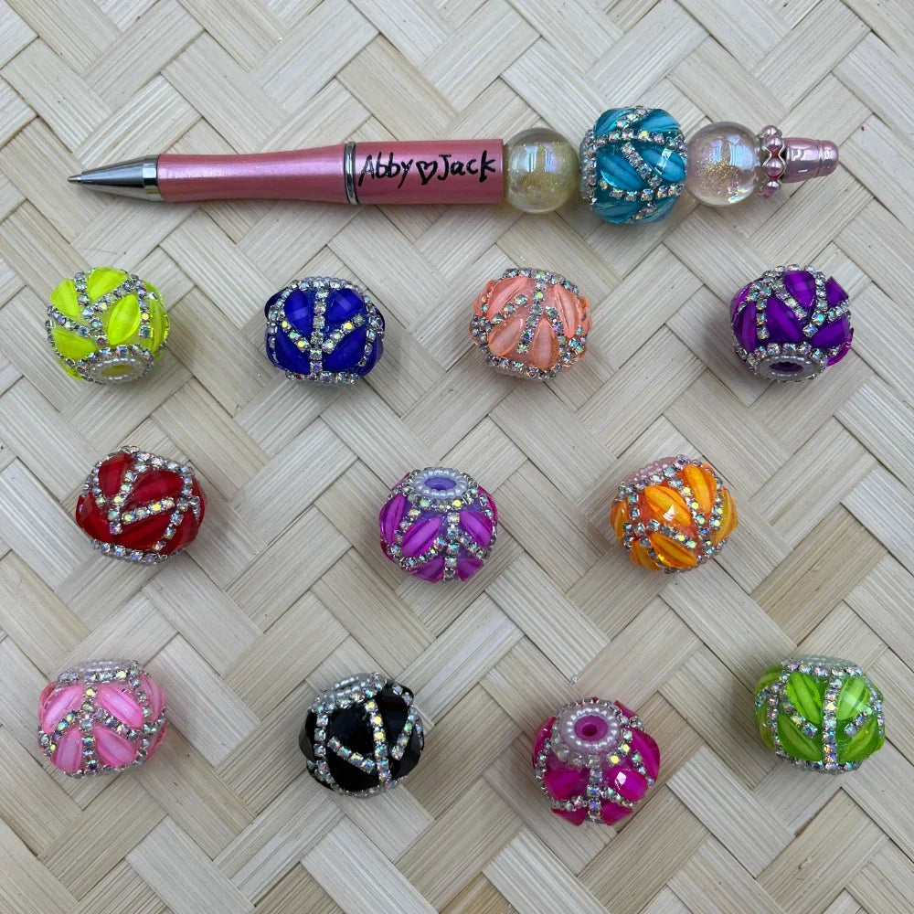 Assorted rhinestone swirl chunky fancy beads in 12 colors including neon yellow, cobalt blue, peach, purple, red, magenta, orange, pink, black, hot pink, and green, displayed on a woven rattan surface alongside a pink beadable pen by Abby Jack.