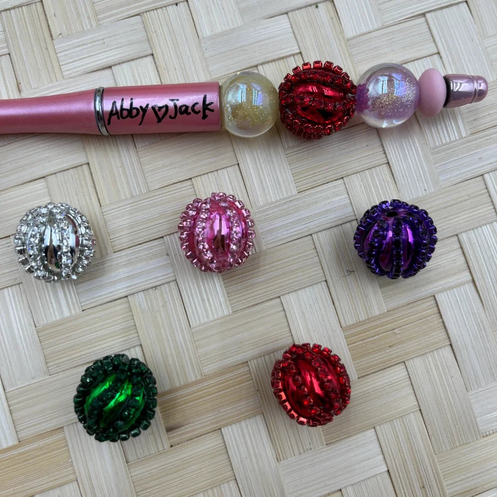 Assorted rhinestone wrapped chunky fancy beads in red, pink, purple, green, and silver displayed on a wicker surface next to a decorated beadable pen labeled Abby Jack.
