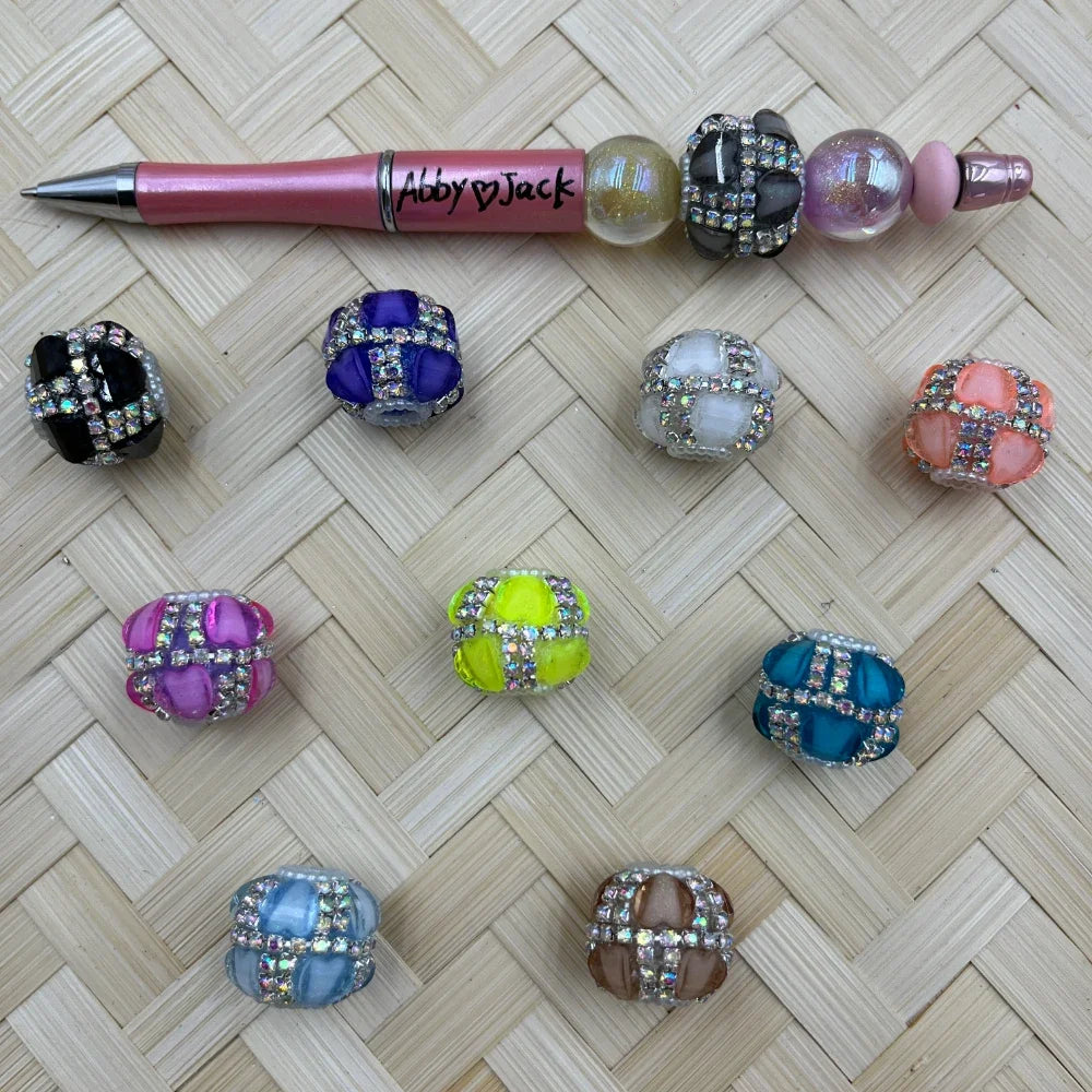 Assorted rhinestone wrapped chunky fancy beads in 9 colors including black, purple, pink, neon yellow, white, orange, teal, blue, and brown displayed on a woven surface next to a decorative pink beadable pen labeled Abby Jack.
