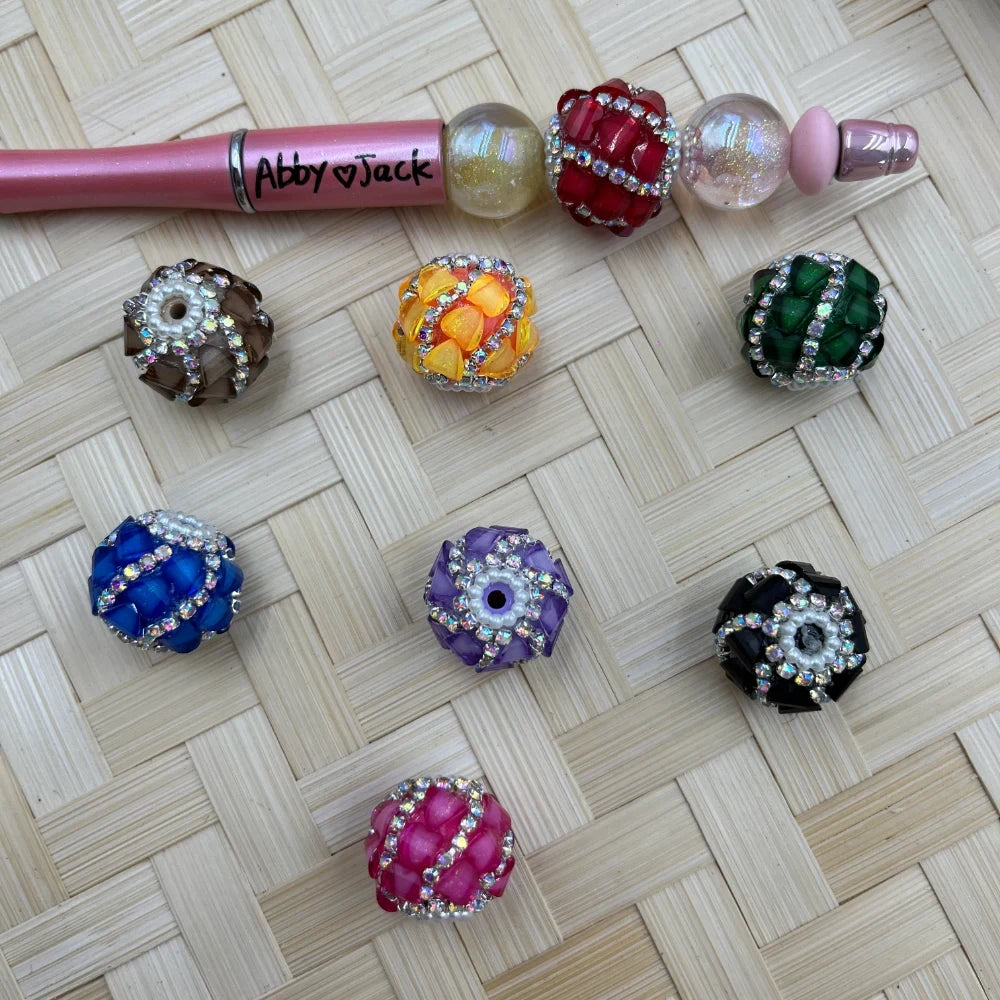 Assorted rhinestone wrapped chunky resin fancy beads in brown, yellow-orange, green, blue, purple, black, and hot pink displayed on a woven surface next to a decorated beadable pen labeled Abby and Jack.