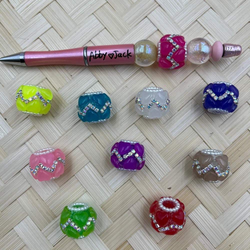 Assorted chunky fancy beads with rhinestone zigzag detailing and pearl edges in 10 colors including hot pink, teal, yellow, purple, red, green, blue, clear, pink, and taupe, displayed alongside a decorative beadable pen on a woven mat.