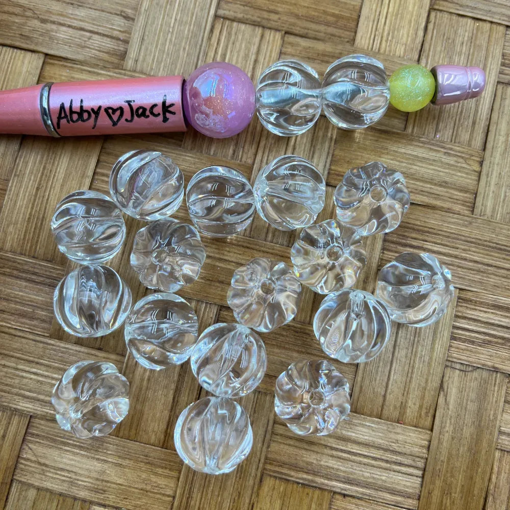 Clear transparent pumpkin-shaped ribbed acrylic beads scattered on a wicker surface next to a pink beadable pen labeled Abby and Jack, showing bead size and texture detail.