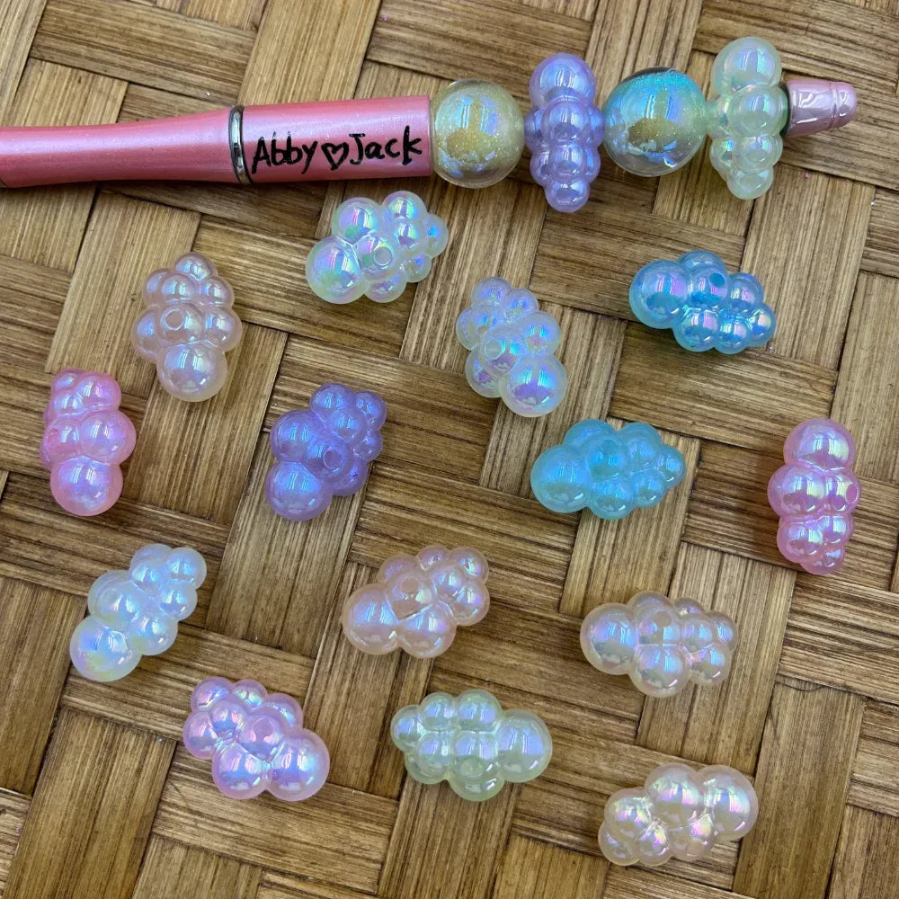 Iridescent AB cloud shape acrylic beads in pastel pink, blue, purple, and clear colors displayed on a wicker surface next to a pink beadable pen branded Abby Jack.