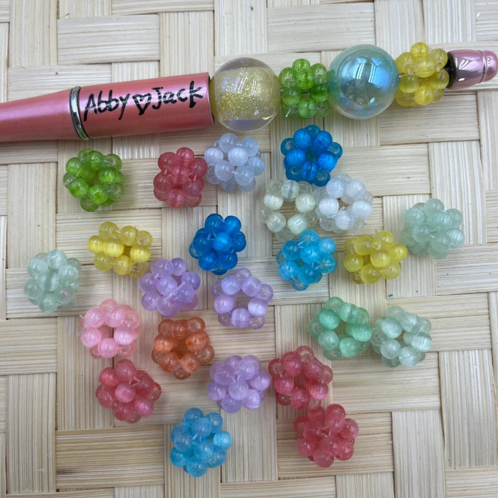 Assorted pastel and bright color cluster woven berry ball fancy beads displayed on a wicker surface next to a pink beadable pen, in colors including pink, blue, green, yellow, purple, teal, and white.