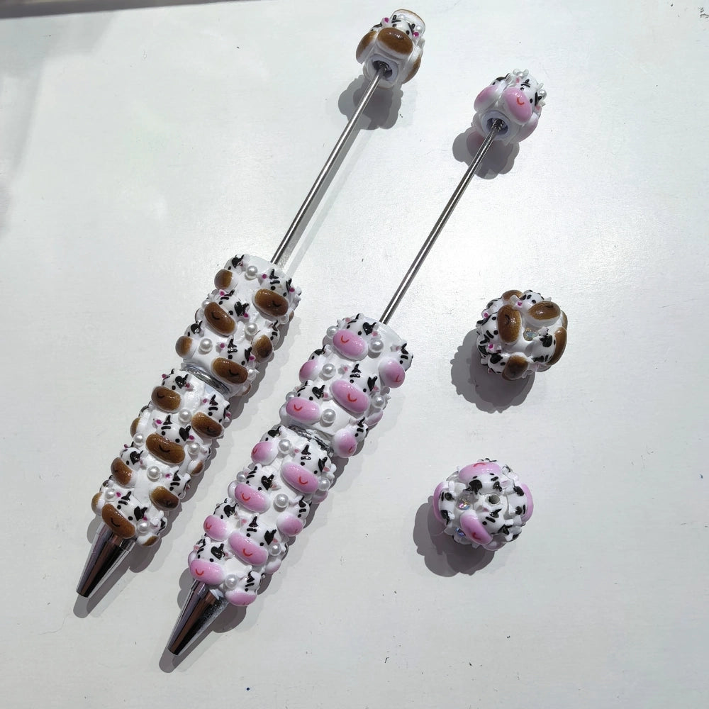 Cow face clay beadable pens with pink noses, brown spots, and pearl accents, shown assembled and with loose beads on white surface.