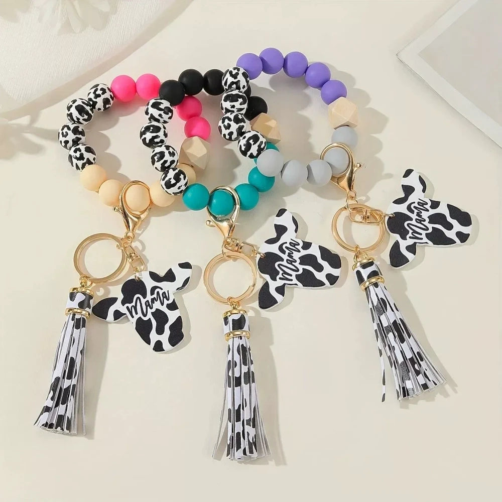 Cow print black and white silicone beads mixed with solid color silicone beads on bracelet keychains with mama butterfly charm and tassel pendants in pink, black, teal, and purple color combinations