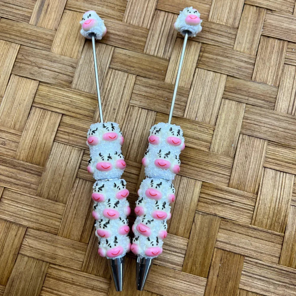 Two white cow print sugar bead pens with pink smiling faces and black spots, featuring glittery sugar-coated beads and silver metal tips, displayed on a woven bamboo background.