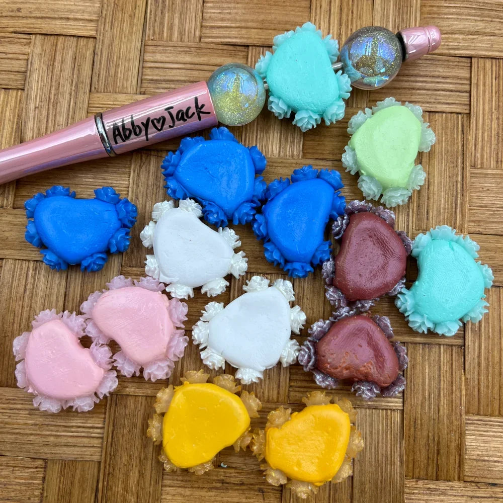 Colorful resin crab shaped fancy beads in blue, white, pink, yellow, teal, green, and maroon colors displayed on a woven surface next to a beadable pen.
