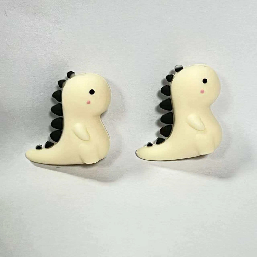 Two cream and black baby dinosaur 3D silicone focal beads with dark spines and pink cheeks, shown from front and side angles on a white background.