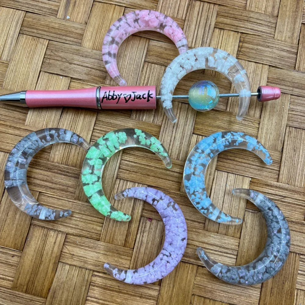 Crescent moon shaped acrylic beads with crushed crystal fill in pink, white, green, blue, purple, and gray on a woven bamboo surface next to a beadable pen.