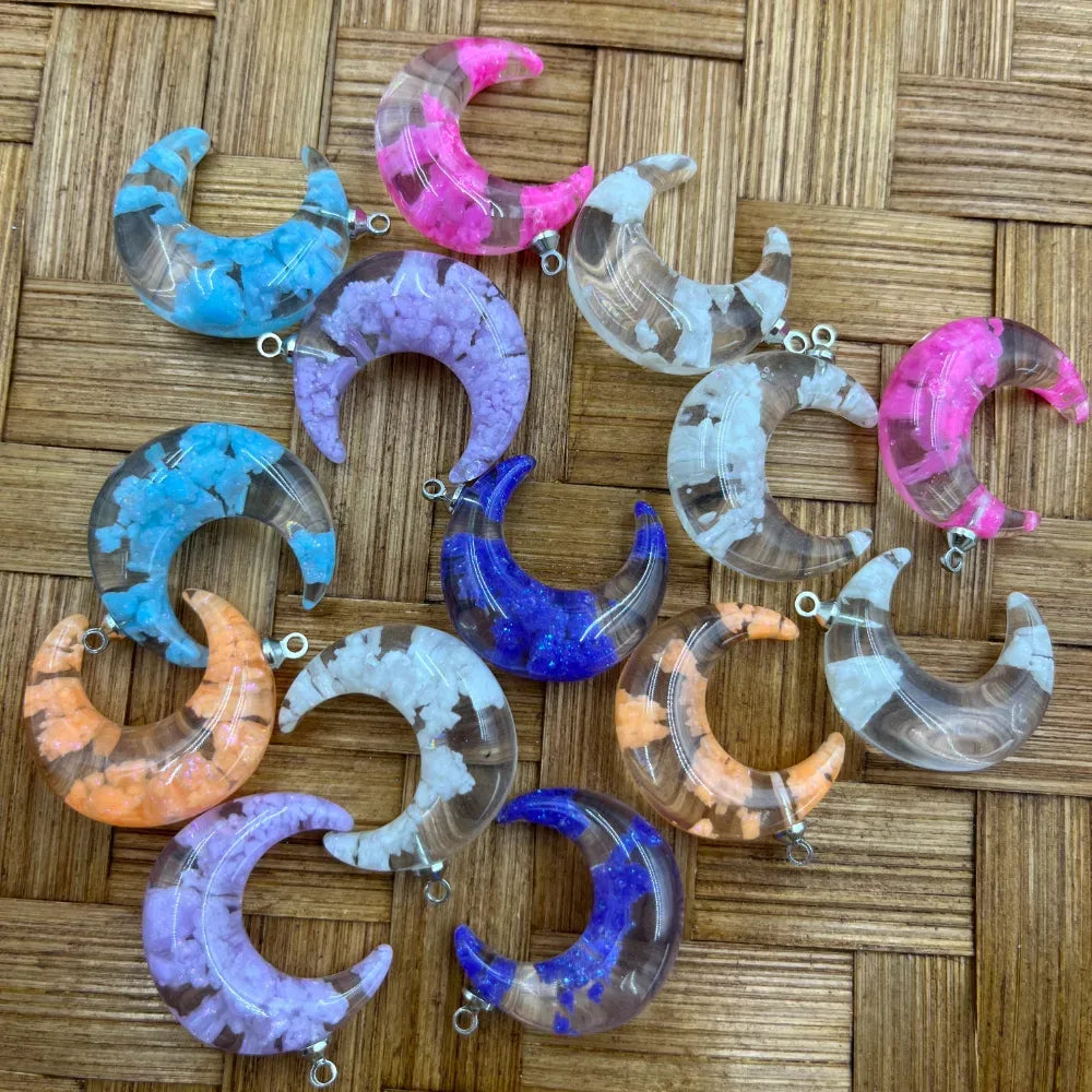 Assorted crescent moon shaped acrylic beads with crushed crystal chip fill in blue, pink, purple, orange, and white on a woven bamboo surface, each with a silver bail loop for jewelry making.