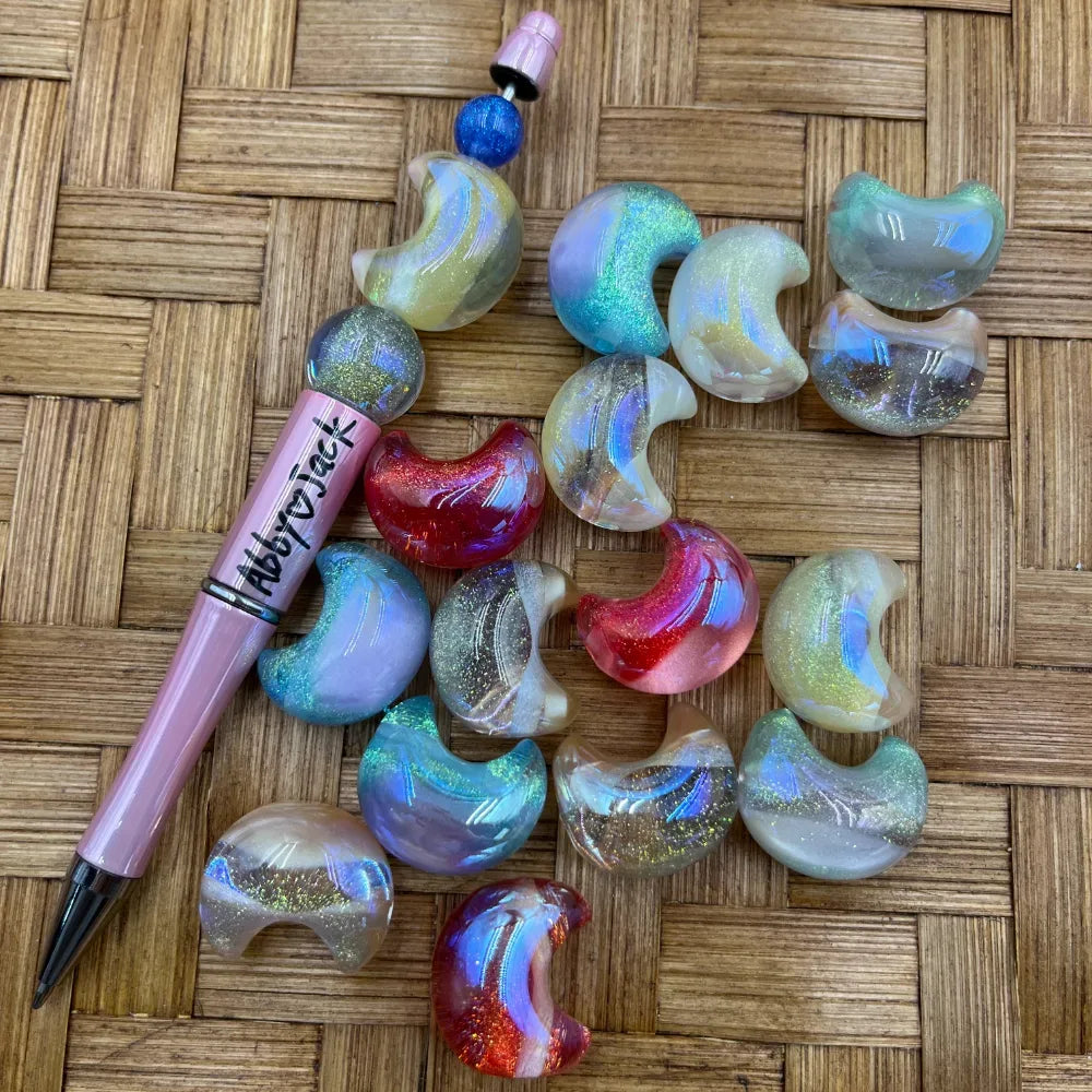 Assorted iridescent glitter crescent moon shaped acrylic beads in pastel and bold colors displayed next to a pink beadable pen on a wicker surface.
