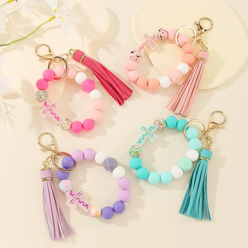 Four colorful silicone bead bracelet keychains with cross charms and leather tassels in pink, hot pink, purple, and teal displayed on a cream background.