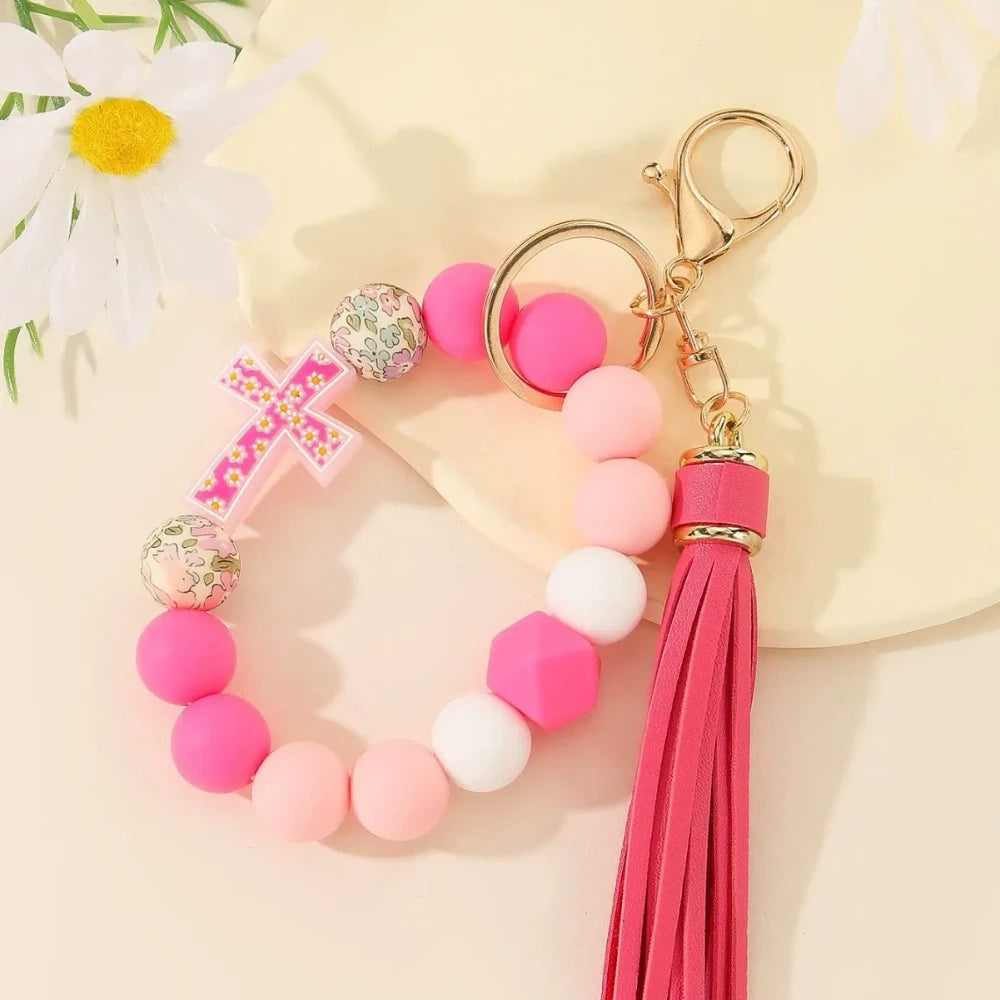 Four colorful silicone bead bracelet keychains with cross charms and leather tassels in pink, hot pink, purple, and teal displayed on a cream background. - A