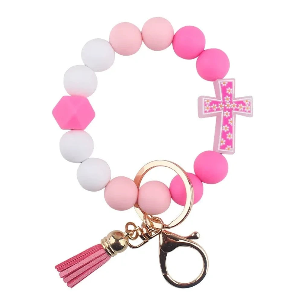 Four colorful silicone bead bracelet keychains with cross charms and leather tassels in pink, hot pink, purple, and teal displayed on a cream background. - B