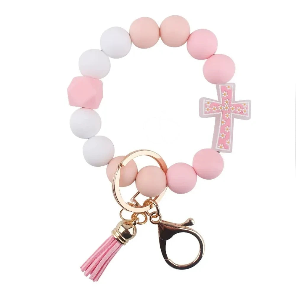 Four colorful silicone bead bracelet keychains with cross charms and leather tassels in pink, hot pink, purple, and teal displayed on a cream background. - C