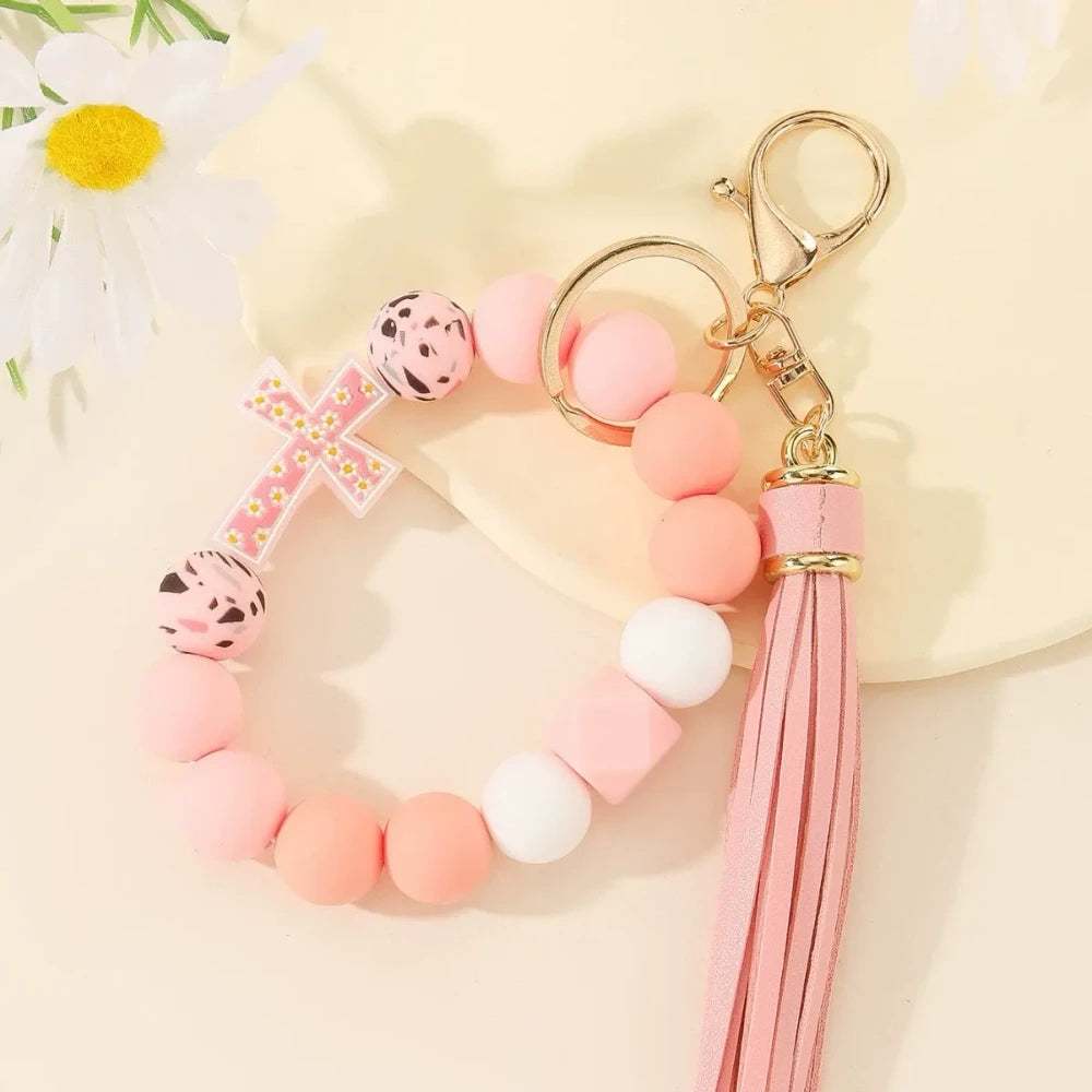Four colorful silicone bead bracelet keychains with cross charms and leather tassels in pink, hot pink, purple, and teal displayed on a cream background. - D