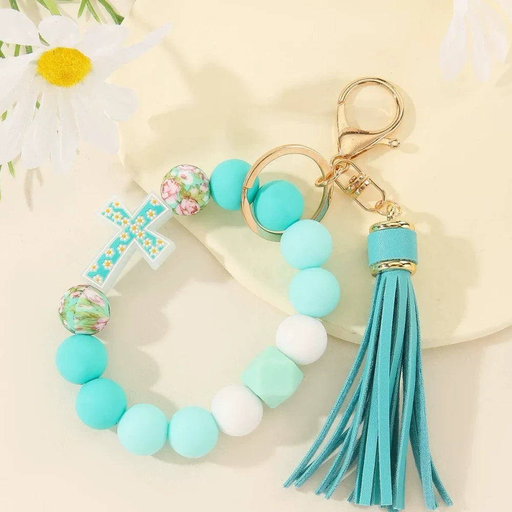 Four colorful silicone bead bracelet keychains with cross charms and leather tassels in pink, hot pink, purple, and teal displayed on a cream background. - E