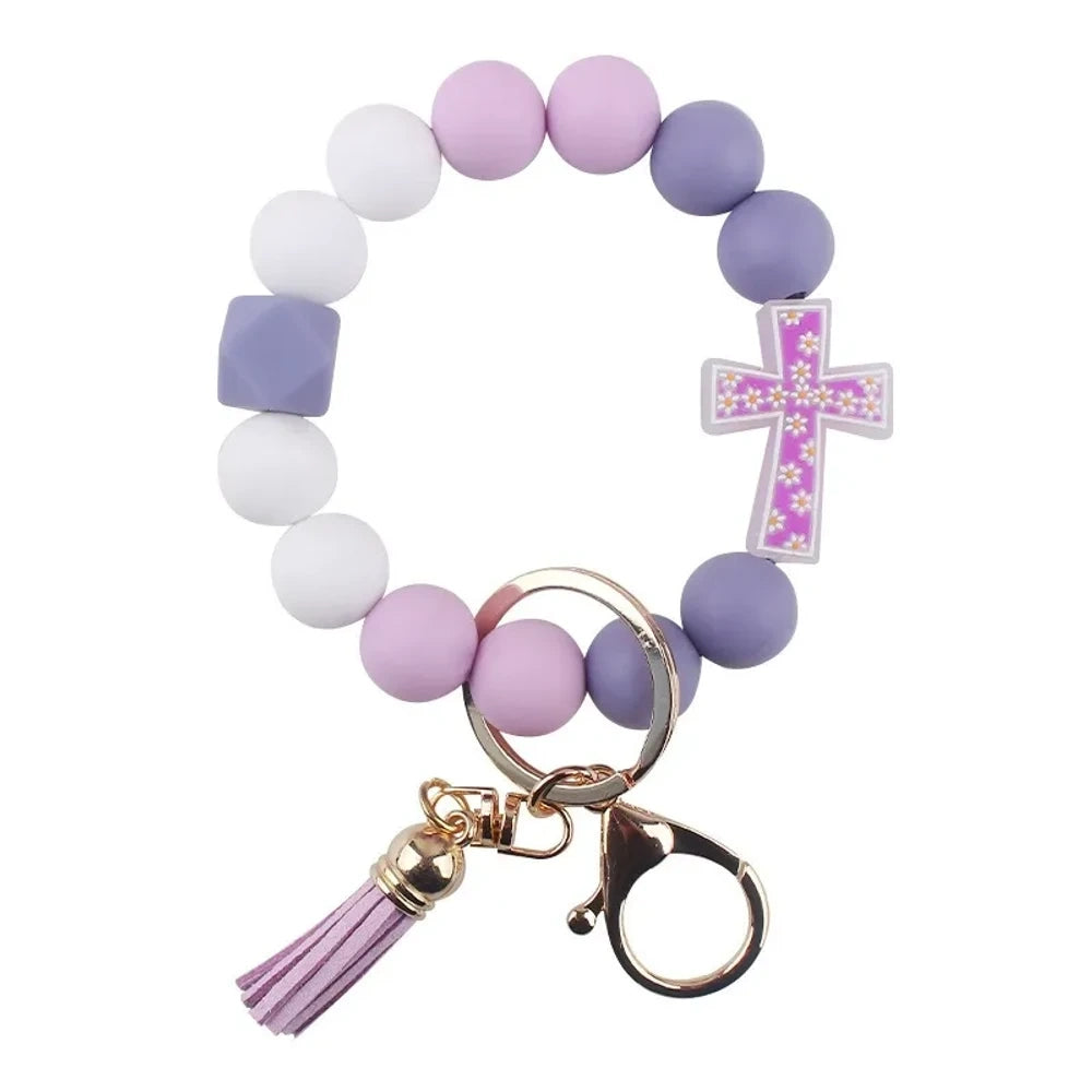 Four colorful silicone bead bracelet keychains with cross charms and leather tassels in pink, hot pink, purple, and teal displayed on a cream background. - F