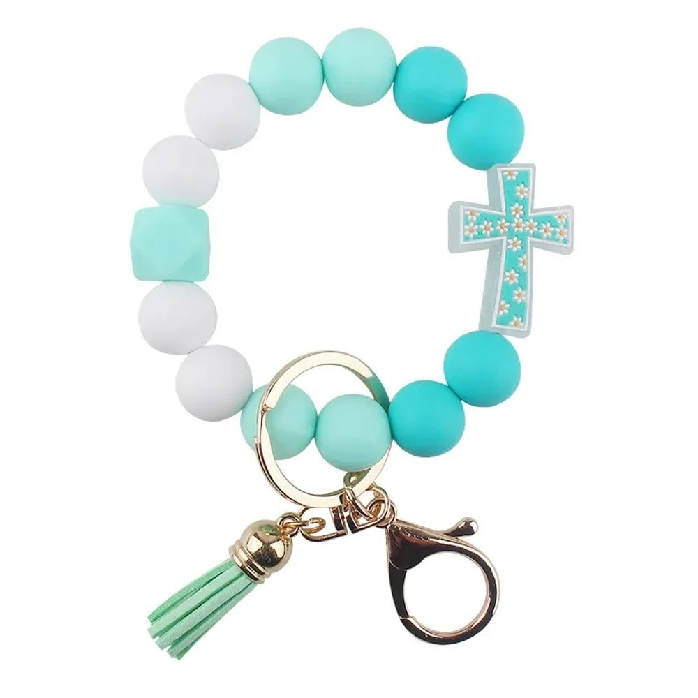 Four colorful silicone bead bracelet keychains with cross charms and leather tassels in pink, hot pink, purple, and teal displayed on a cream background. - G