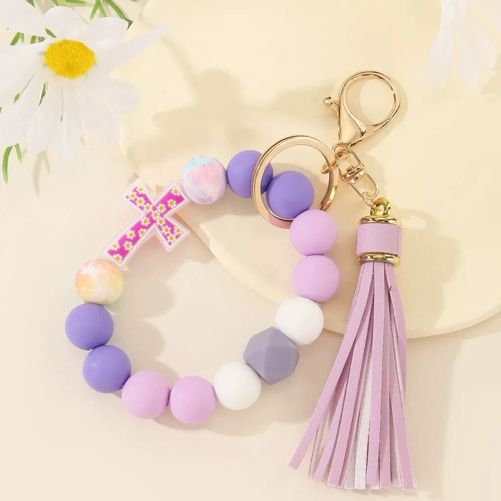 Four colorful silicone bead bracelet keychains with cross charms and leather tassels in pink, hot pink, purple, and teal displayed on a cream background. - H