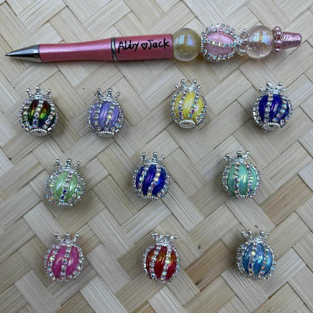 Nine colorful crown rhinestone ladybug fancy beads in assorted enamel colors including purple, yellow, blue, green, pink, red, and teal, displayed on a woven bamboo surface next to a pink beadable pen labeled Abby and Jack.