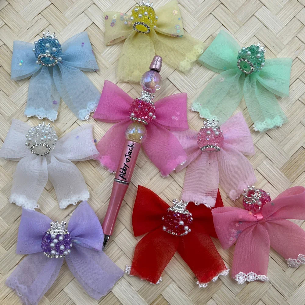 Assorted colorful crown rhinestone and pearl cluster fancy beads displayed with decorative organza bows and a pink beadable pen labeled Abby Jack, shown in blue, yellow, green, white, purple, red, and pink on a woven bamboo background.