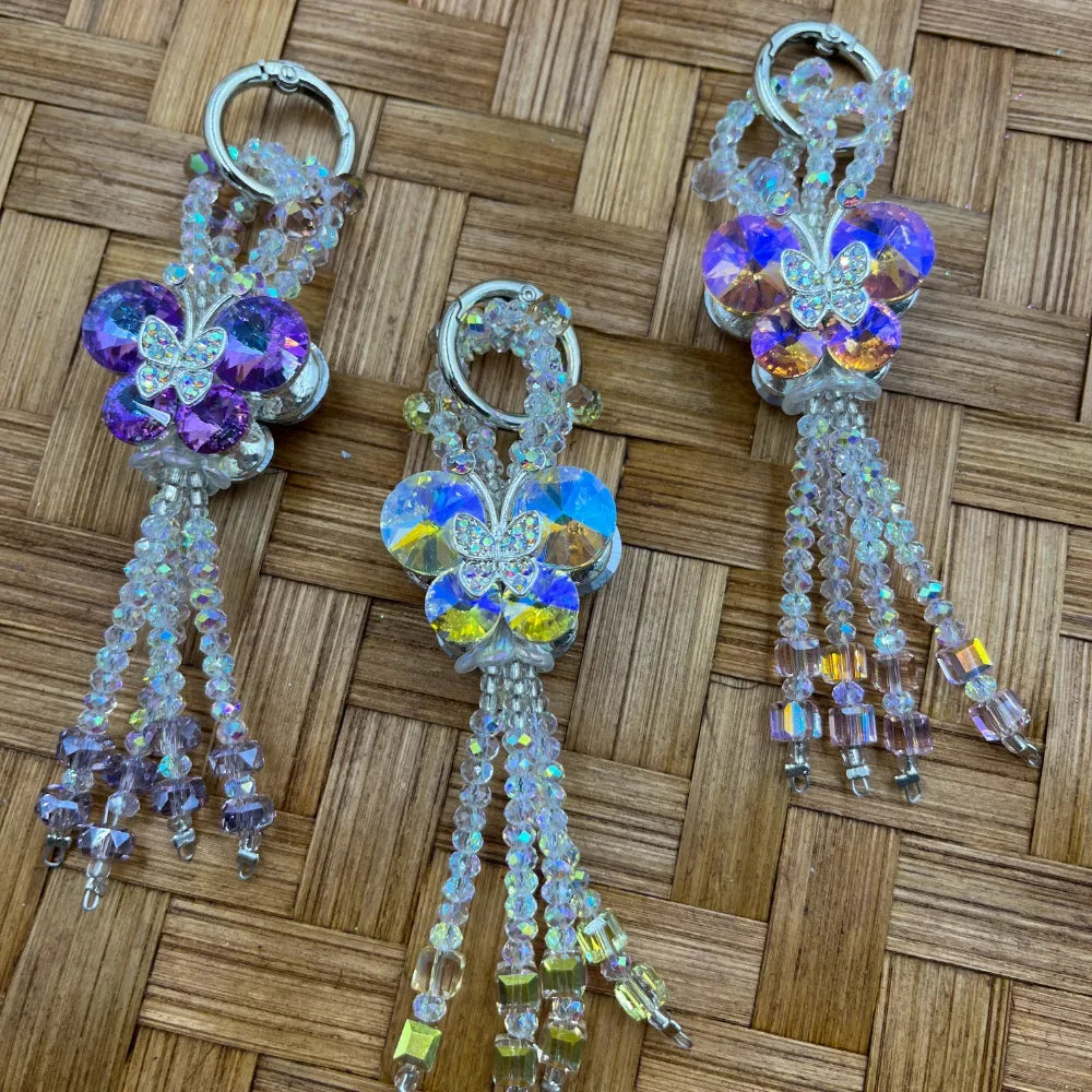 Three crystal AB butterfly flower beadable keychains with iridescent tassel bead strands in purple, aurora borealis, and lavender pink on a woven bamboo surface