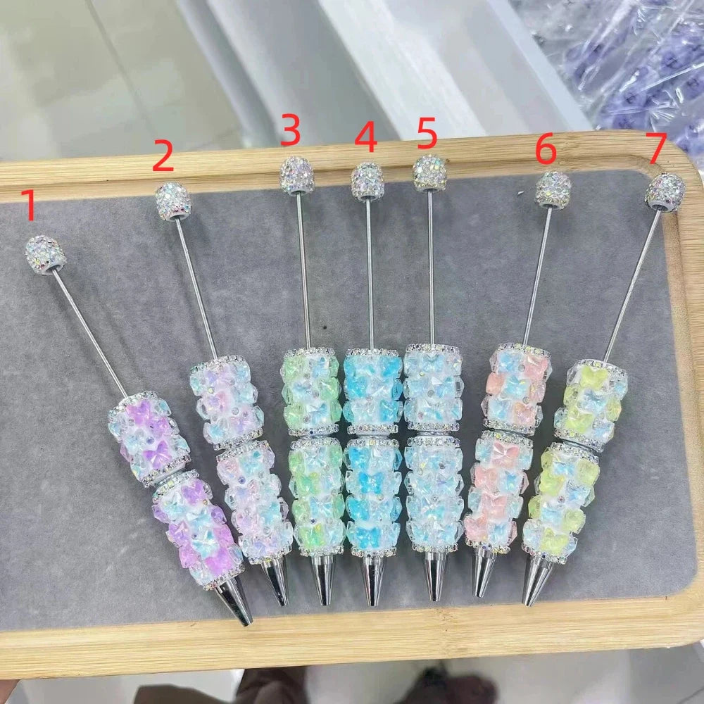 Set of 7 beadable pens decorated with pastel crystal bear charms and rhinestone spacers, topped with sparkly rhinestone balls, arranged on a wooden tray numbered 1 through 7 in colors purple, blue-green, mint green, sky blue, light blue, pink, and yellow-green.