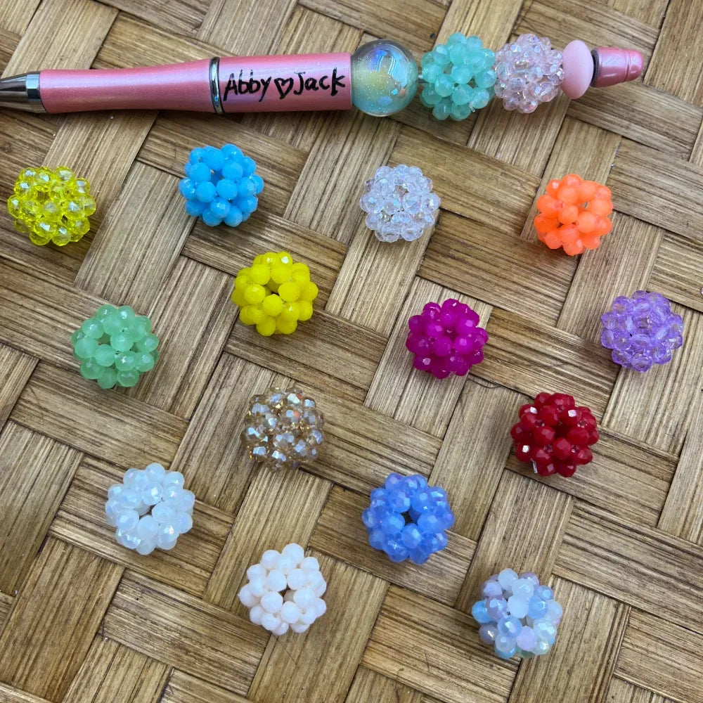 Assorted colorful crystal berry cluster ball fancy beads in yellow, blue, green, white, magenta, orange, purple, red, and clear displayed on a woven surface next to a pink beadable pen labeled Abby and Jack.