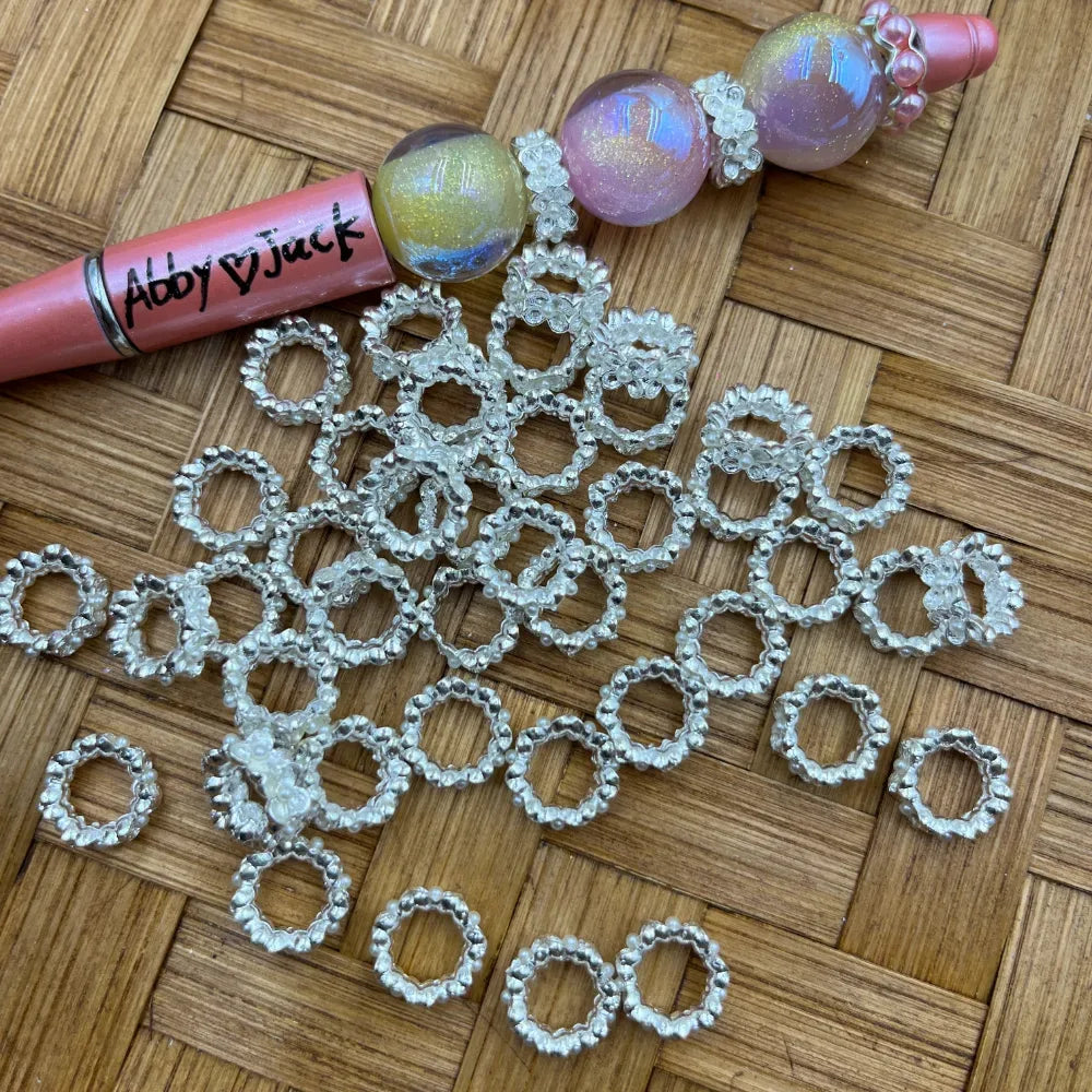Crystal clear and silver beaded ring metal spacers arranged on a woven surface next to a decorated beadable pen, showing various sizes of circular beaded rings for DIY jewelry making.