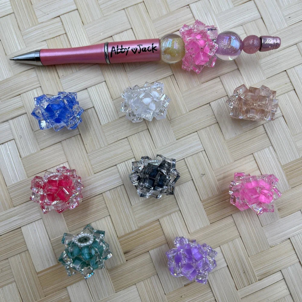 Assorted crystal cluster flower fancy beads in blue, clear, pink, red, black, teal, purple, and brown colors displayed on woven surface with a decorated beadable pen