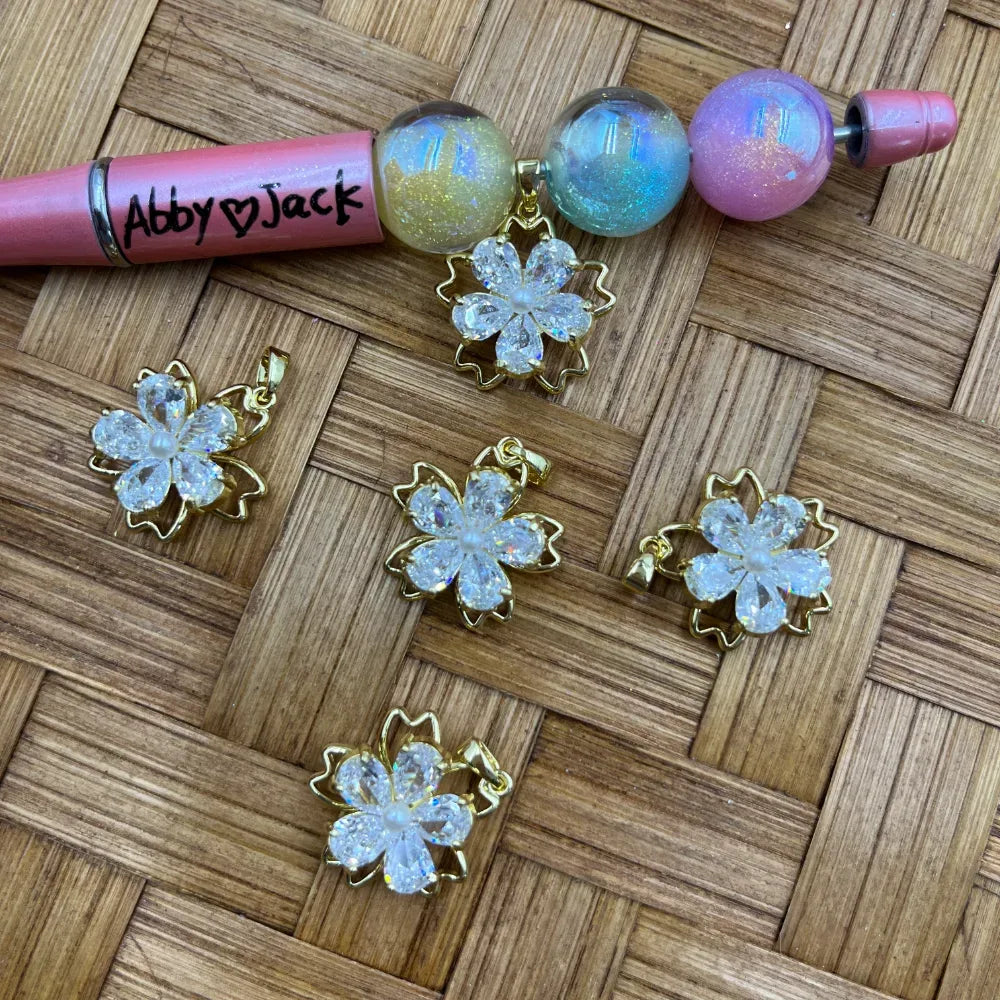 Five crystal CZ sakura cherry blossom flower charms with gold bail settings displayed on woven bamboo surface next to a pink beadable pen with iridescent globe beads.