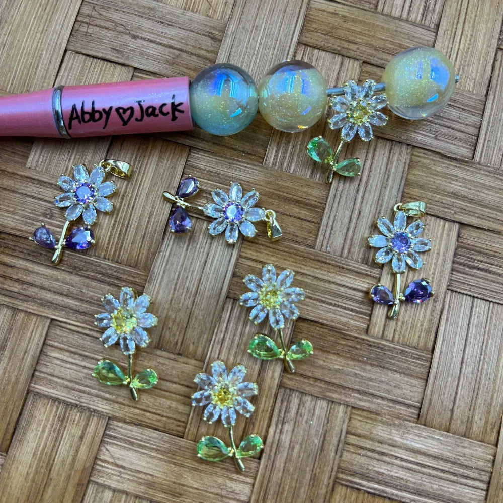 Assorted crystal daisy flower charm bail beads with purple and green gemstone accents alongside iridescent opal globe bail beads on a woven bamboo surface, next to a pink beadable pen labeled Abby loves Jack.