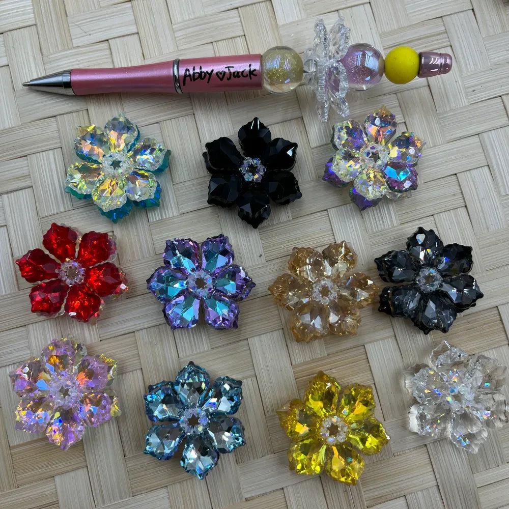 11 crystal flower cluster fancy beads in assorted colors including red, black, purple, teal, yellow, clear, and iridescent AB, displayed on wicker surface next to a beadable pen.