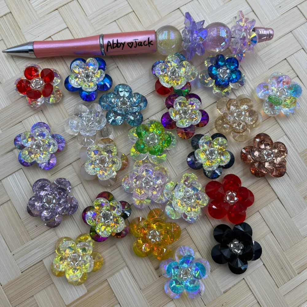 Assorted crystal flower cluster fancy beads in multiple colors including red, blue, purple, yellow, black, and iridescent AB finish, displayed on woven bamboo surface next to a pink beadable pen.
