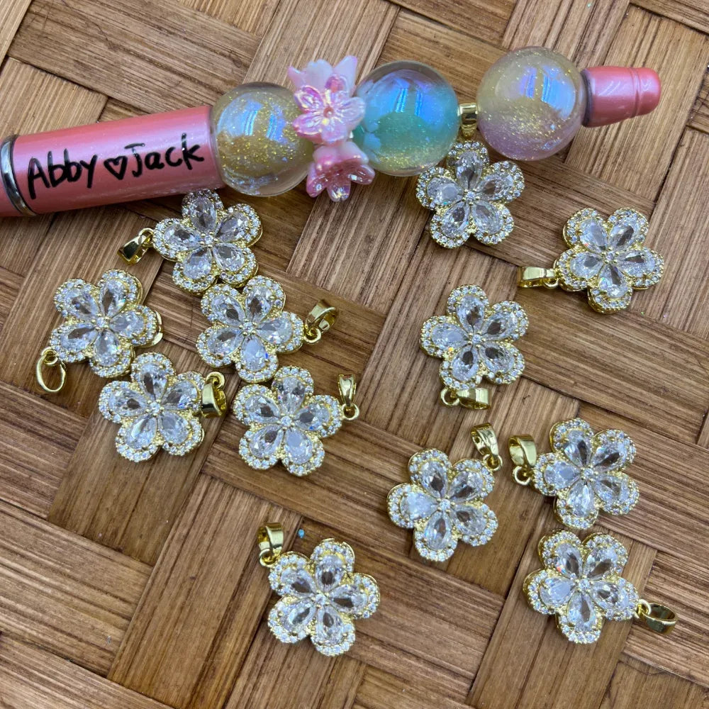 Crystal clear CZ rhinestone flower-shaped gold bail pendants arranged on a woven surface, shown with decorative beadable pens for scale.