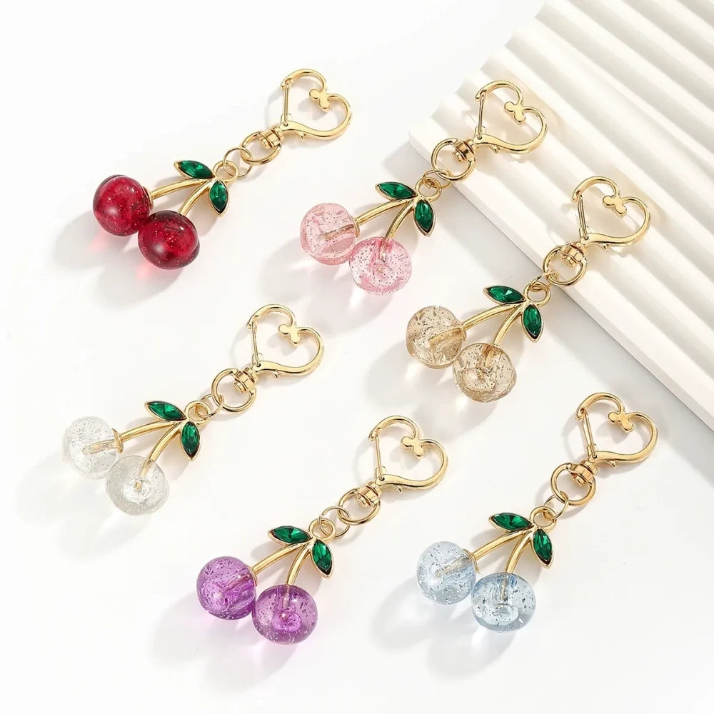 Six crystal glitter cherry charm keychains in red, pink, clear, champagne, purple, and blue with gold heart-shaped lobster clasps and green enamel leaves on white background.