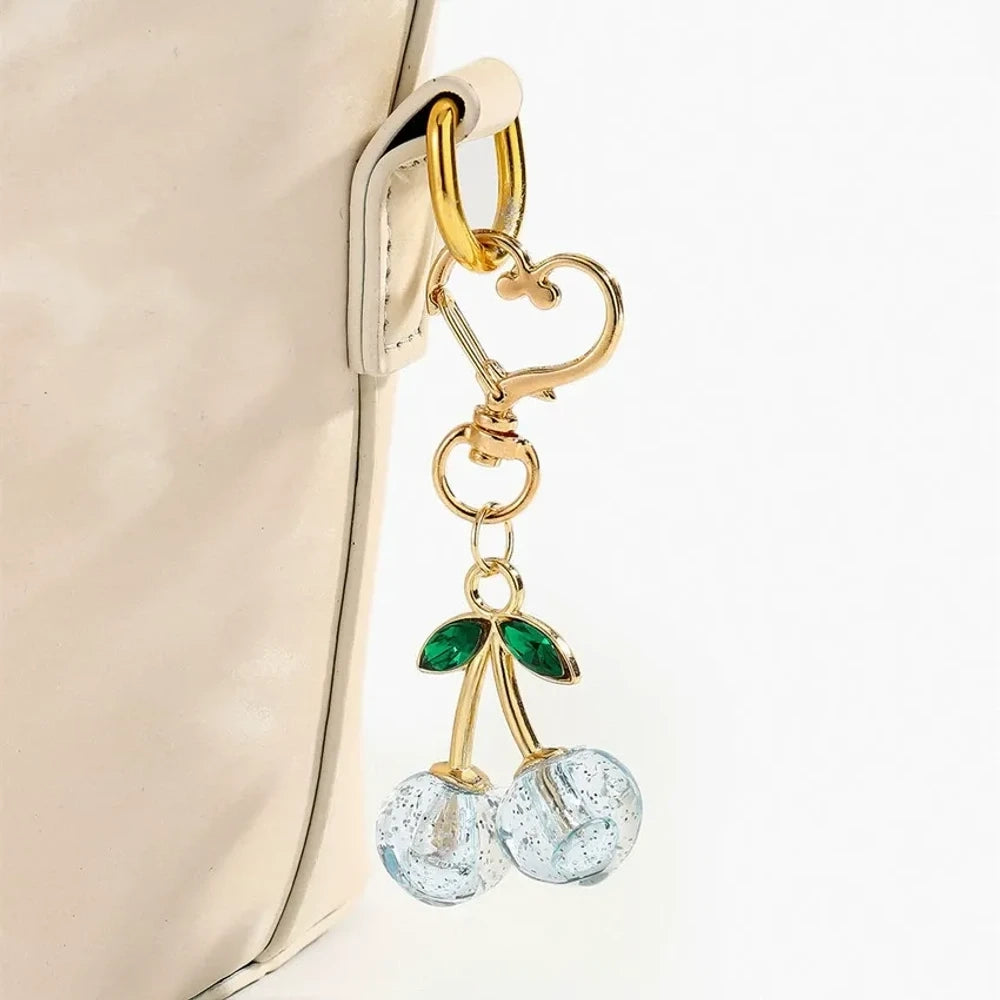 Six crystal glitter cherry charm keychains in red, pink, clear, champagne, purple, and blue with gold heart-shaped lobster clasps and green enamel leaves on white background. - Blue